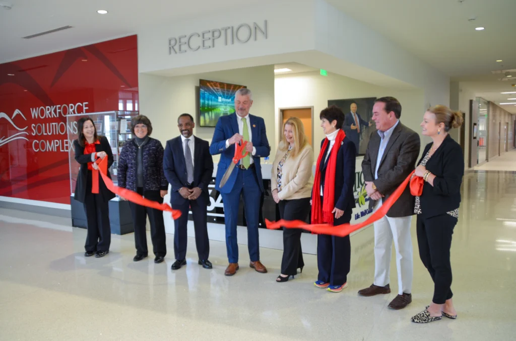 A ribbon-cutting ceremony at the Workforce Solutions Complex shows a group of formally dressed individuals smiling as a man in a blue suit and green tie prepares to cut a red ribbon with oversized scissors. The word “RECEPTION” is visible above the desk behind them.