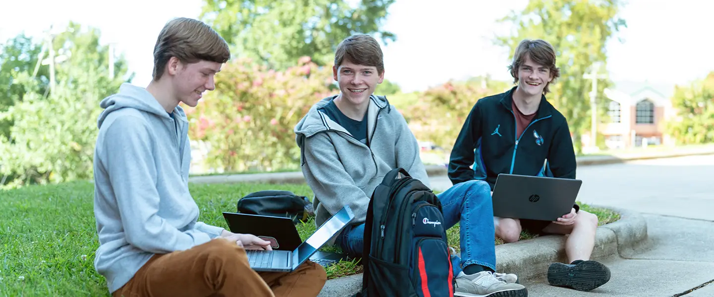 Three students sitting outside using laptops and talking together.