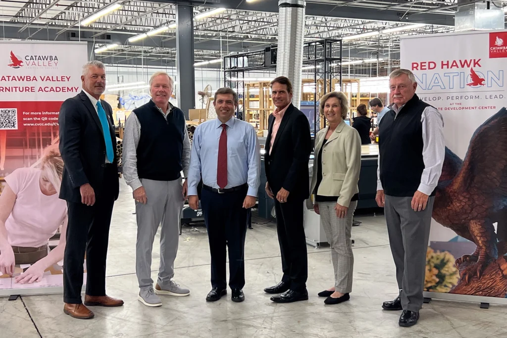 Seven individuals stand in a line inside a modern industrial facility, posing in front of banners for the Catawba Valley Furniture Academy and Red Hawk Nation. The group includes both men and women in business attire, with manufacturing equipment visible in the background.