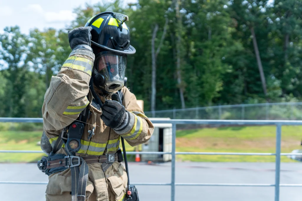 A firefighter in full gear, including a helmet and breathing mask, adjusts their equipment outdoors near a training facility with trees in the background.