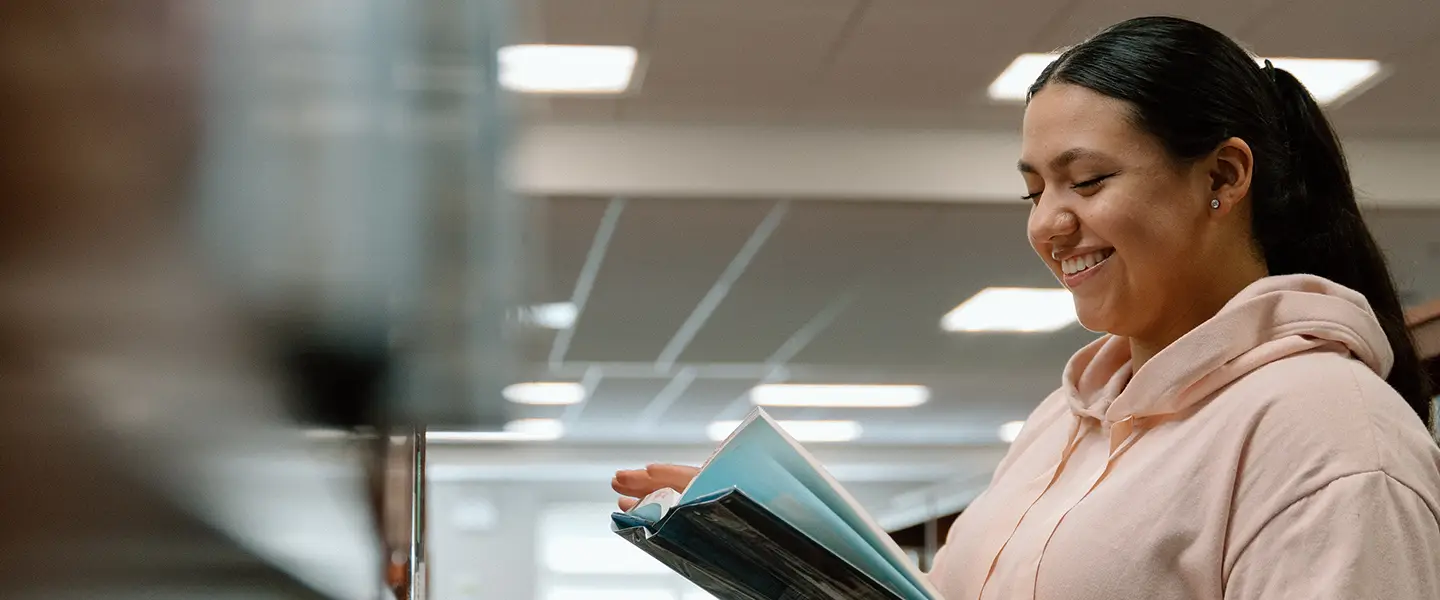 Student browsing a shelf in the library, holding an open book.