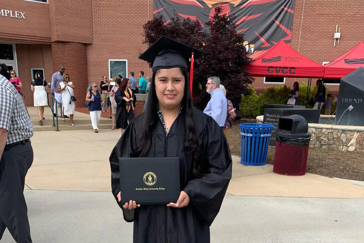 A young woman in a black graduation cap and gown smiles while holding her diploma in front of a brick campus building. Behind her, friends and family gather to celebrate, with a red "CVCC" tent and a banner displaying a red hawk logo in the background.