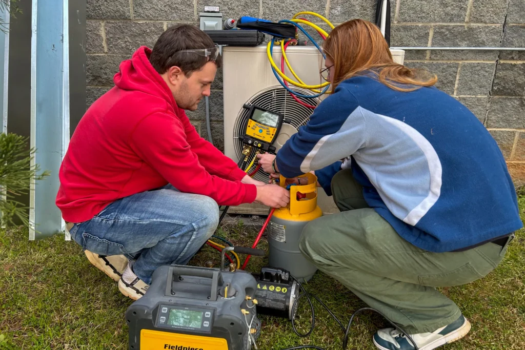 Two HVAC students crouch on the grass outside a building, working together on an air conditioning unit. They are using gauges and hoses to test or recharge the system, with various tools and equipment laid out nearby. One student wears a red hoodie and safety goggles; the other wears a blue and gray jacket.