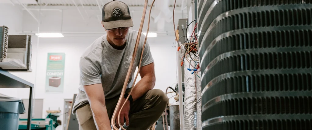 A technician wearing a gray shirt, khaki pants, safety glasses, and a dark baseball cap is working with copper tubing near an HVAC unit in a well-lit workshop. He is crouched beside the equipment, focusing on the connections. Exposed wires and tools are visible around the workspace, with a large vented component of the HVAC system dominating the right side of the image. A poster with the brand “Bryant” is visible in the background.