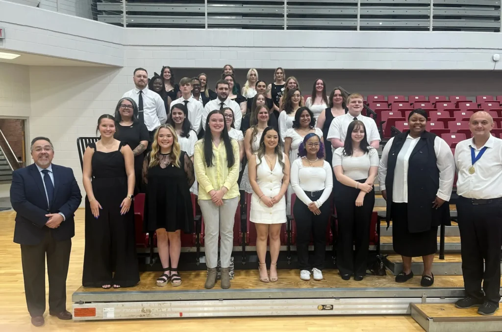 A large group of students and faculty members pose together in a gymnasium. The group is dressed in formal black and white attire, with some variations including a yellow shirt and a gold medal. They are arranged in several rows on bleachers, smiling at the camera. The setting includes red bleacher seats and a wood-floored gym.