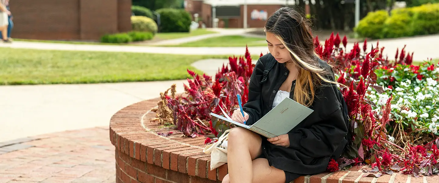 Female student sitting in vibrant and floral campus quad writing in a notebook.