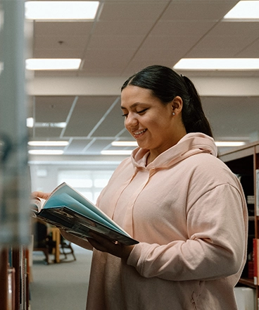 A smiling student wearing a light pink hoodie reads a book while standing between bookshelves in a library. She holds the book open with both hands and appears engaged with the content. The setting is bright and organized, with overhead lights and additional study spaces visible in the background.