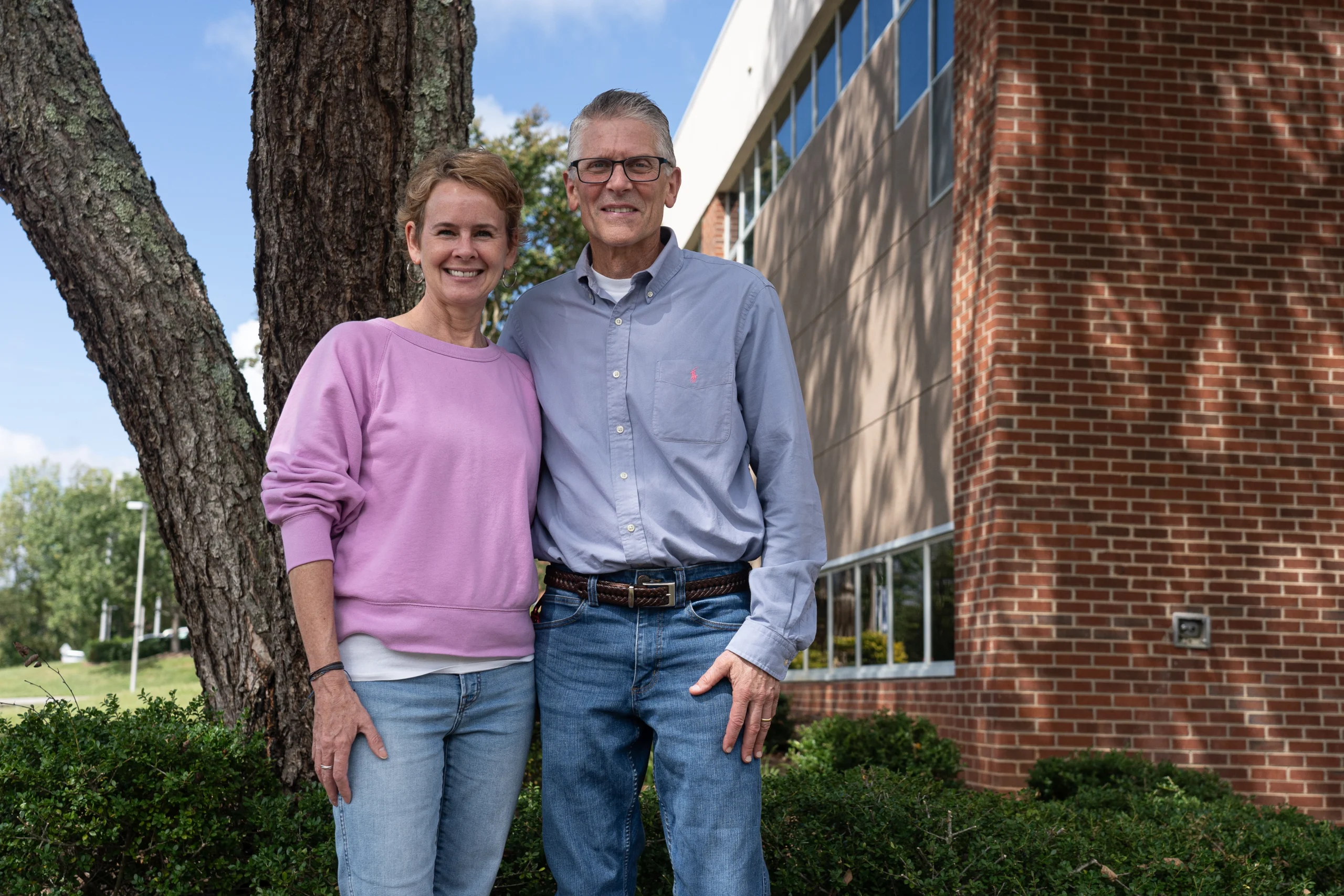 Two adults stand outdoors smiling, with one wearing a light purple sweatshirt and the other in a light blue button-down shirt. They are positioned next to a tree and in front of a brick and glass building on a sunny day.