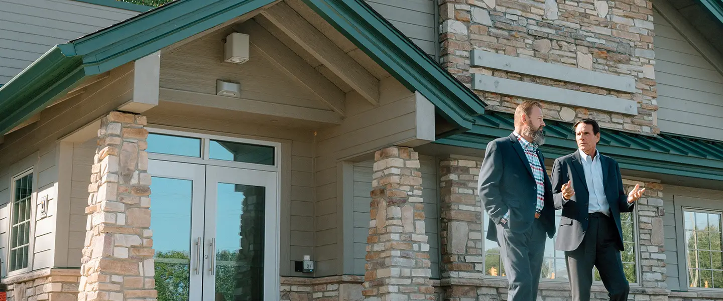 Two professionals in business attire talking outside a stone-accented building.