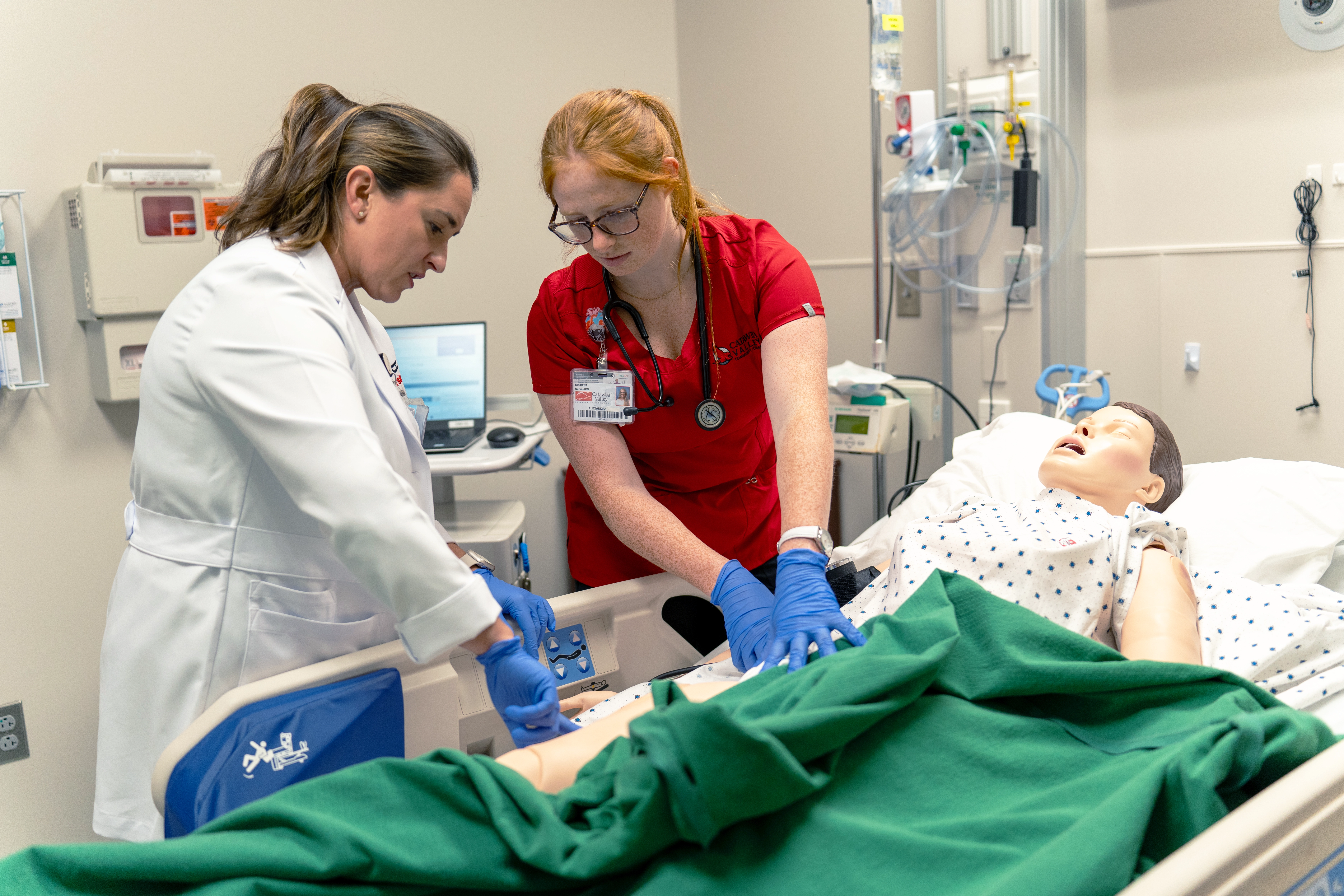 A nursing student in red scrubs and an instructor in a white coat work together during a medical simulation on a patient manikin in a hospital bed. The student performs chest compressions while the instructor monitors and assists. Medical equipment and a computer are visible in the background.