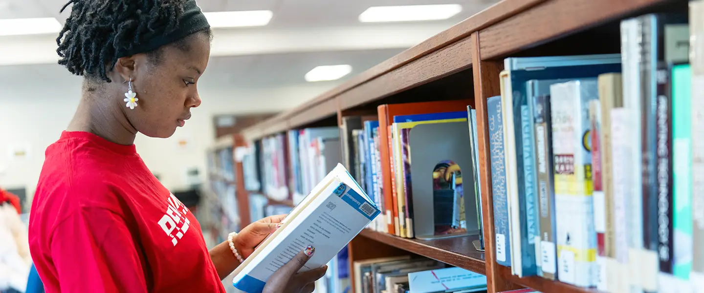 Student browsing a shelf in the library, holding an open book.