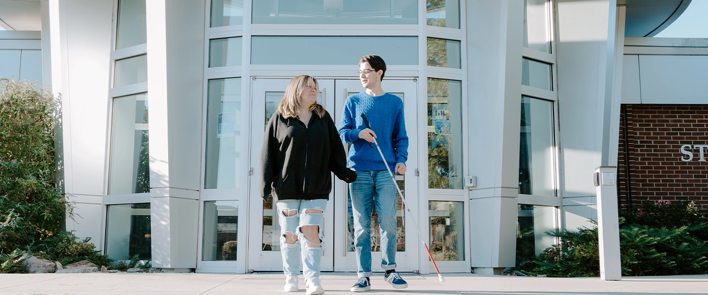 Two students walk together in front of a modern building with large glass windows and white framing at Catawba Valley Community College. One student, wearing a blue sweater and holding a white cane, appears to be visually impaired. The other student wears a black hoodie and ripped jeans, walking supportively beside him. The background includes landscaped greenery and part of a brick wall with the word “STUDENT” partially visible. The atmosphere is inclusive and friendly.