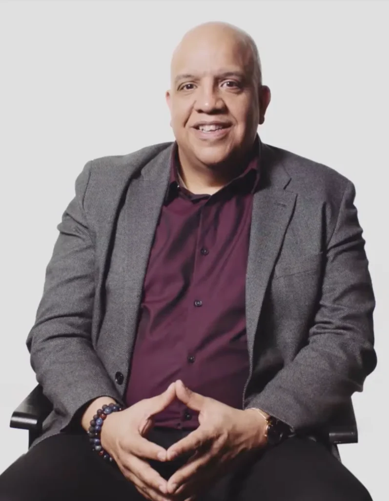 A bald man in a gray blazer and maroon shirt is sitting in a chair against a plain white background. He is smiling and has his hands loosely clasped together in front of him. He wears a watch and beaded bracelets, giving a calm and confident demeanor.