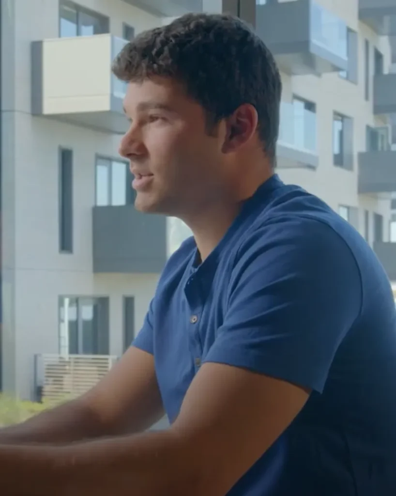 A man in a blue shirt is sitting indoors near a large window, speaking or listening attentively. Behind him, modern apartment buildings with balconies are visible through the window. The setting appears to be a casual or professional conversation space.