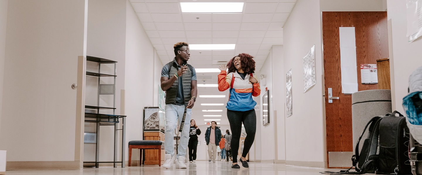 Two students walk down a brightly lit hallway at Catawba Valley Community College, smiling and engaged in conversation. One wears a gray vest and carries a lanyard, while the other wears a colorful windbreaker jacket. Backpacks and a rolled-up poster are visible on the floor to the right, and several other students walk in the background. The hallway has beige walls, white ceiling tiles, and classroom doors along both sides.