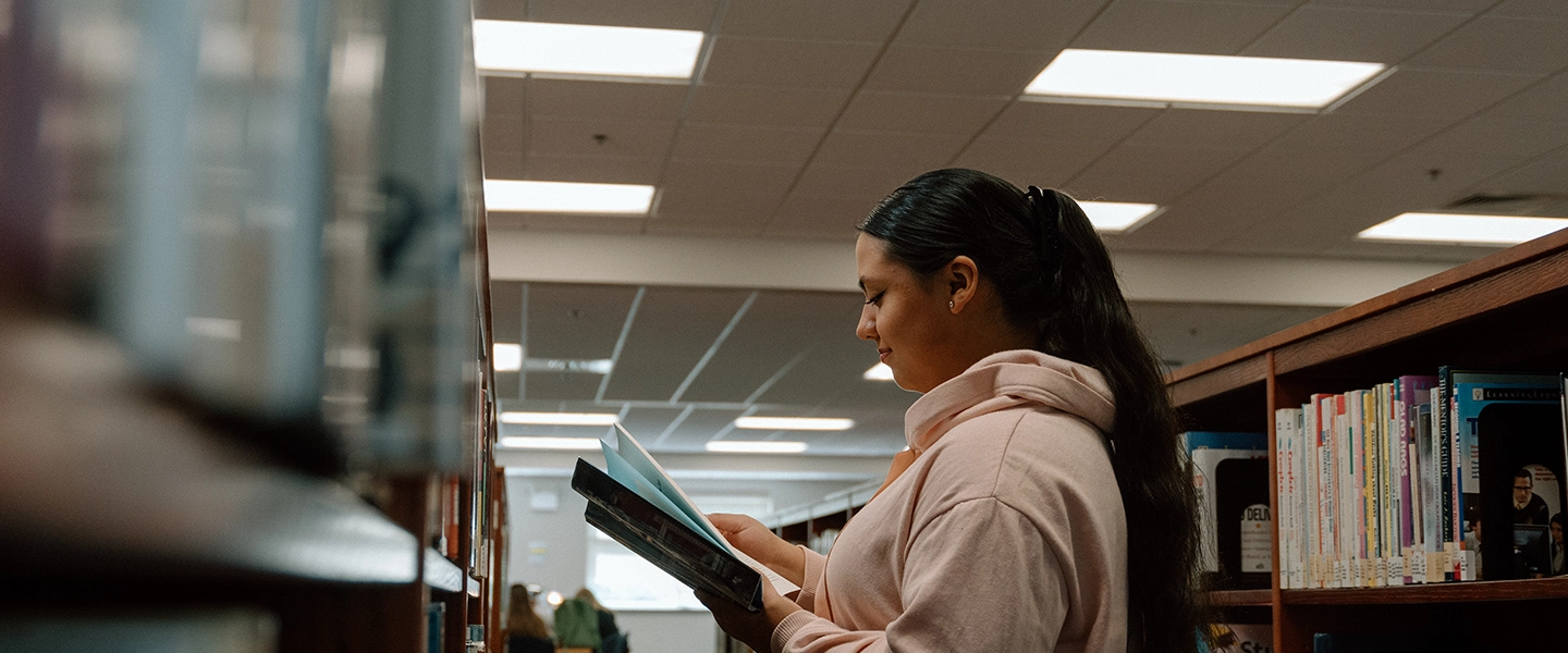 A student stands between bookshelves in a library or resource center at Catawba Valley Community College, reading a book. She wears a light pink hoodie and has long, dark hair pulled back in a ponytail. The space is brightly lit with overhead lights, and rows of books are neatly arranged on the shelves around her. The atmosphere is quiet and focused.