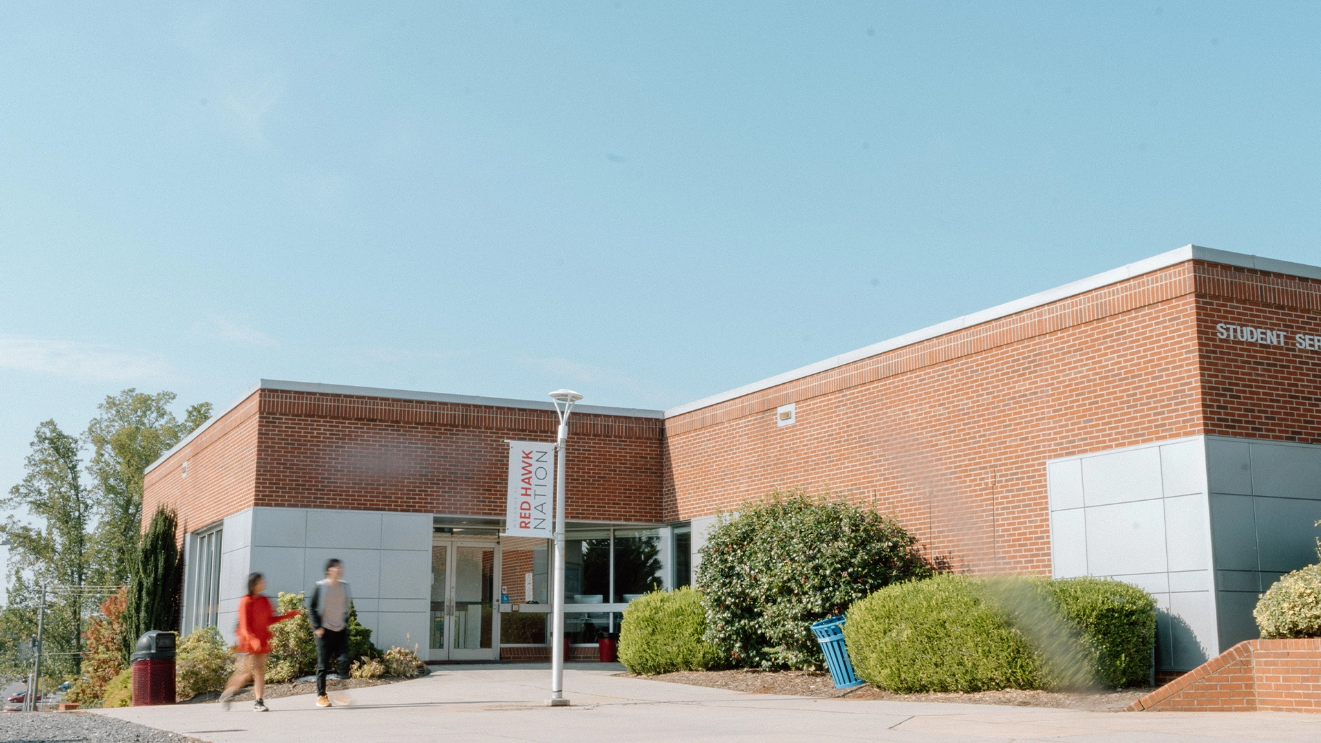 Photo of the Student Services building at Catawba Valley Community College on a clear, sunny day. The structure features red brick walls with silver-gray paneling near the entrance. A banner reading “Red Hawk Nation” hangs from a nearby lamp post. Two blurred students are walking past the entrance, and the area is surrounded by landscaped bushes and trees. The sky is bright blue with minimal clouds.