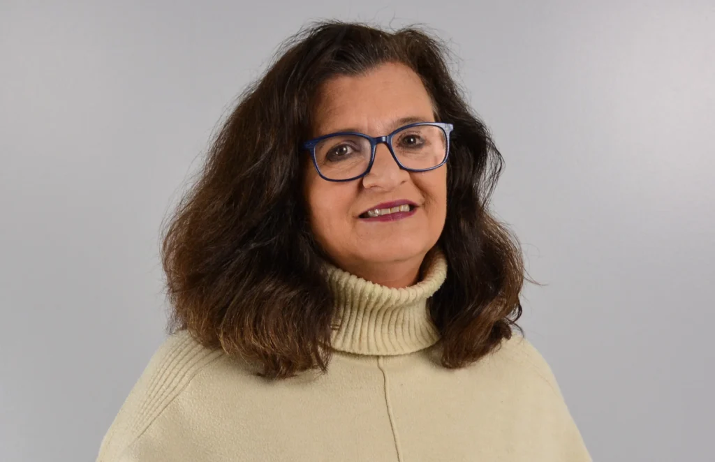Professional portrait of a woman with long dark brown hair wearing blue-framed glasses and a cream-colored turtleneck sweater. She is smiling softly and posed against a plain gray background.
