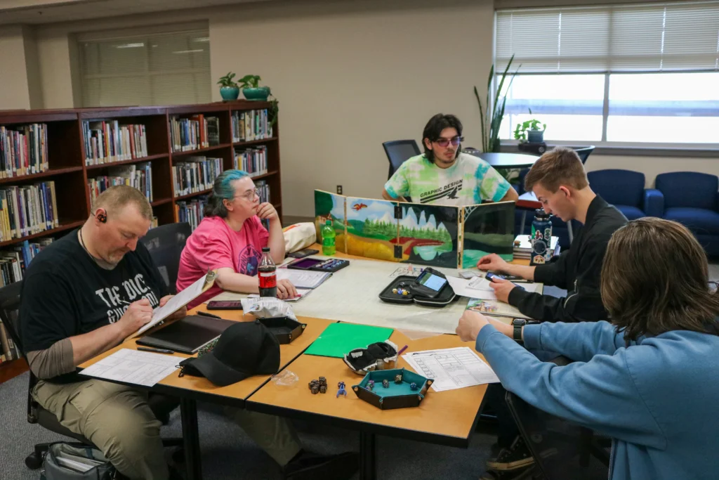 Members of the Dungeons and Dragons club sit around a table playing a tabletop role‑playing game with character sheets, dice, and a dungeon master screen.