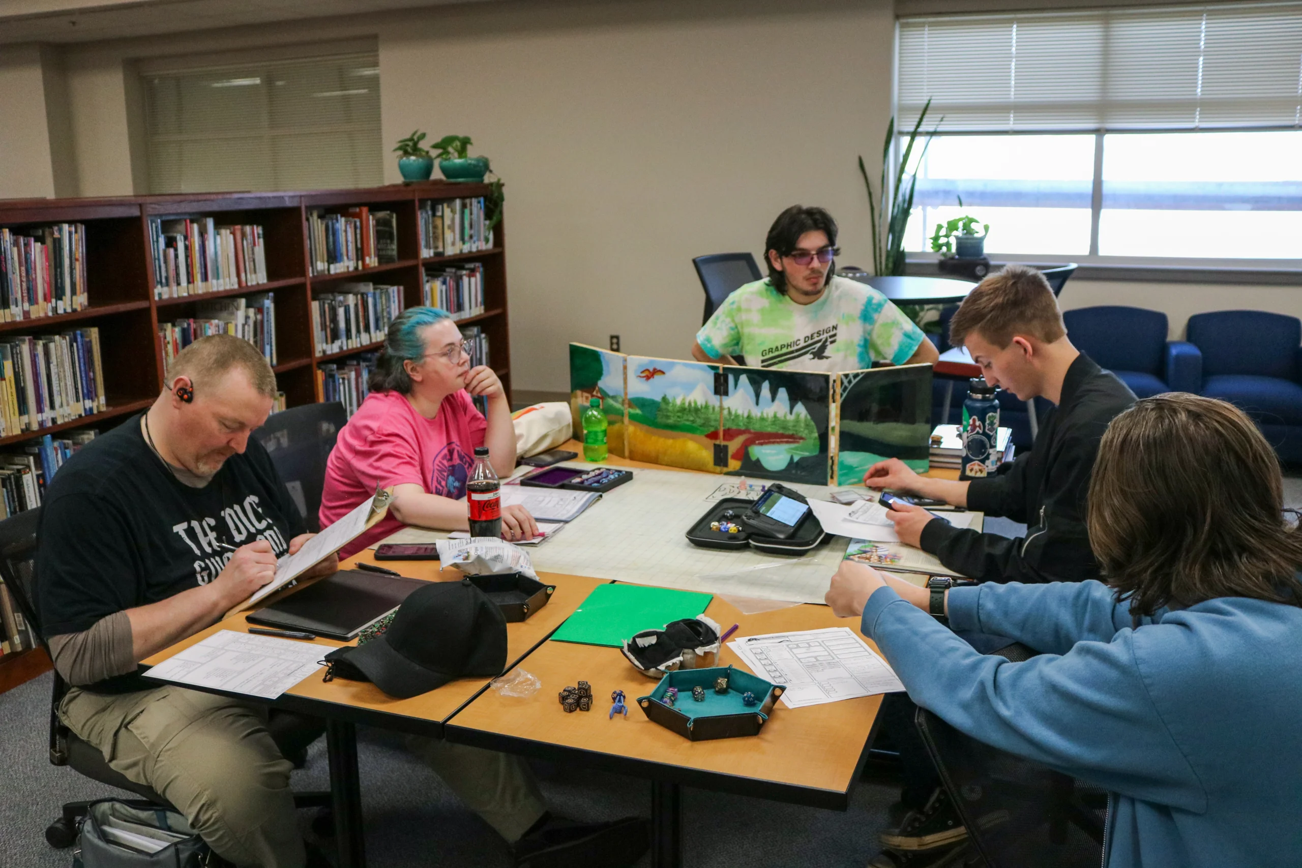 Members of the Dungeons and Dragons club sit around a table playing a tabletop role‑playing game with character sheets, dice, and a dungeon master screen.