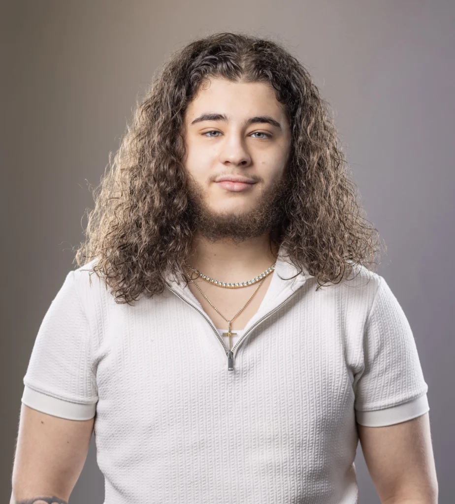 CVCC alum Nic J. stands centered in a studio portrait with shoulder‑length curly hair and facial hair, wearing a light zip‑collar short‑sleeve shirt and layered necklaces, photographed against a soft gray studio backdrop.