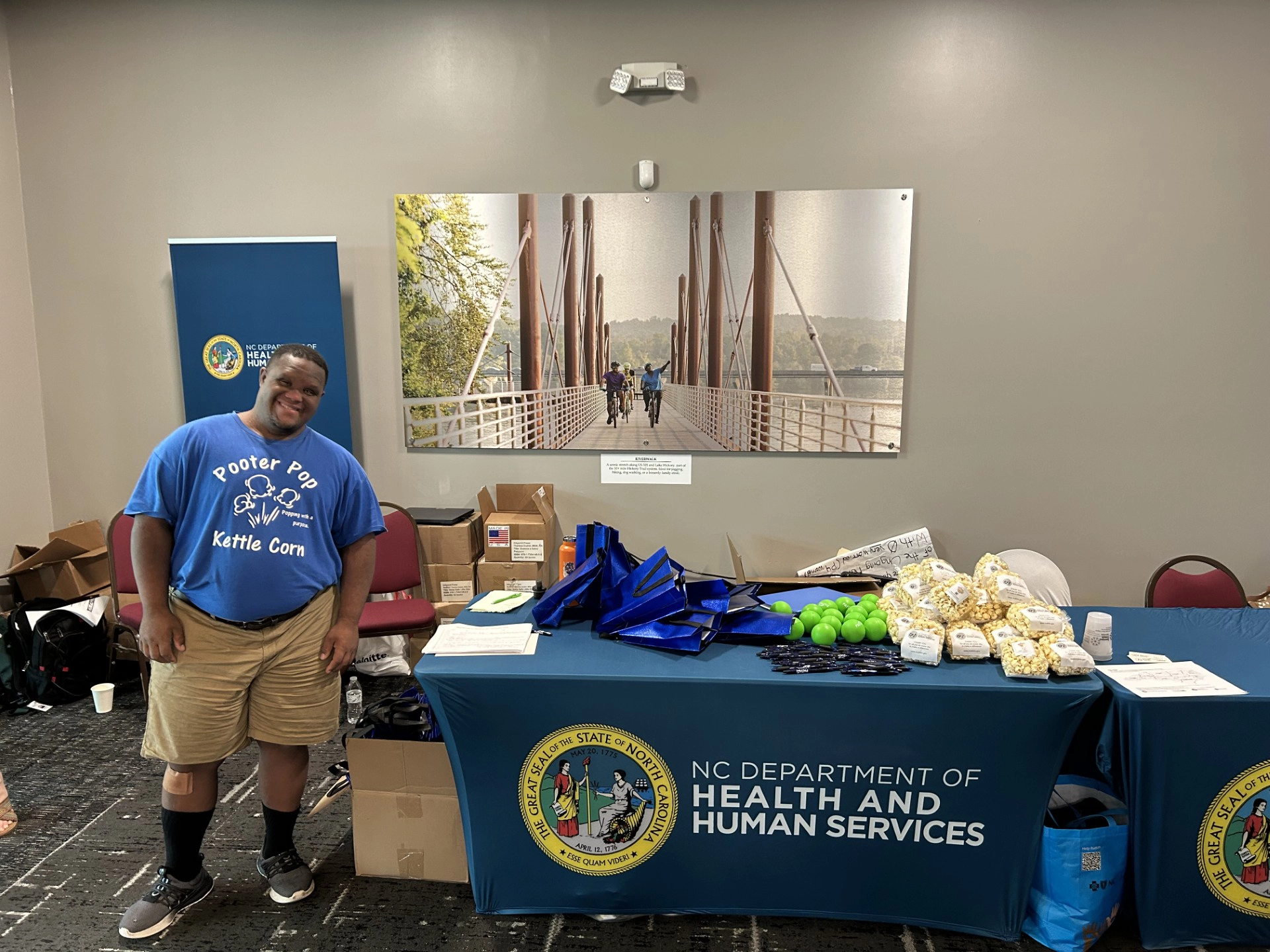 Malcolm from Pooter Pop stands beside an event table branded with the NC Department of Health and Human Services logo, displaying packaged kettle corn, tote bags, and promotional items in an indoor event space.