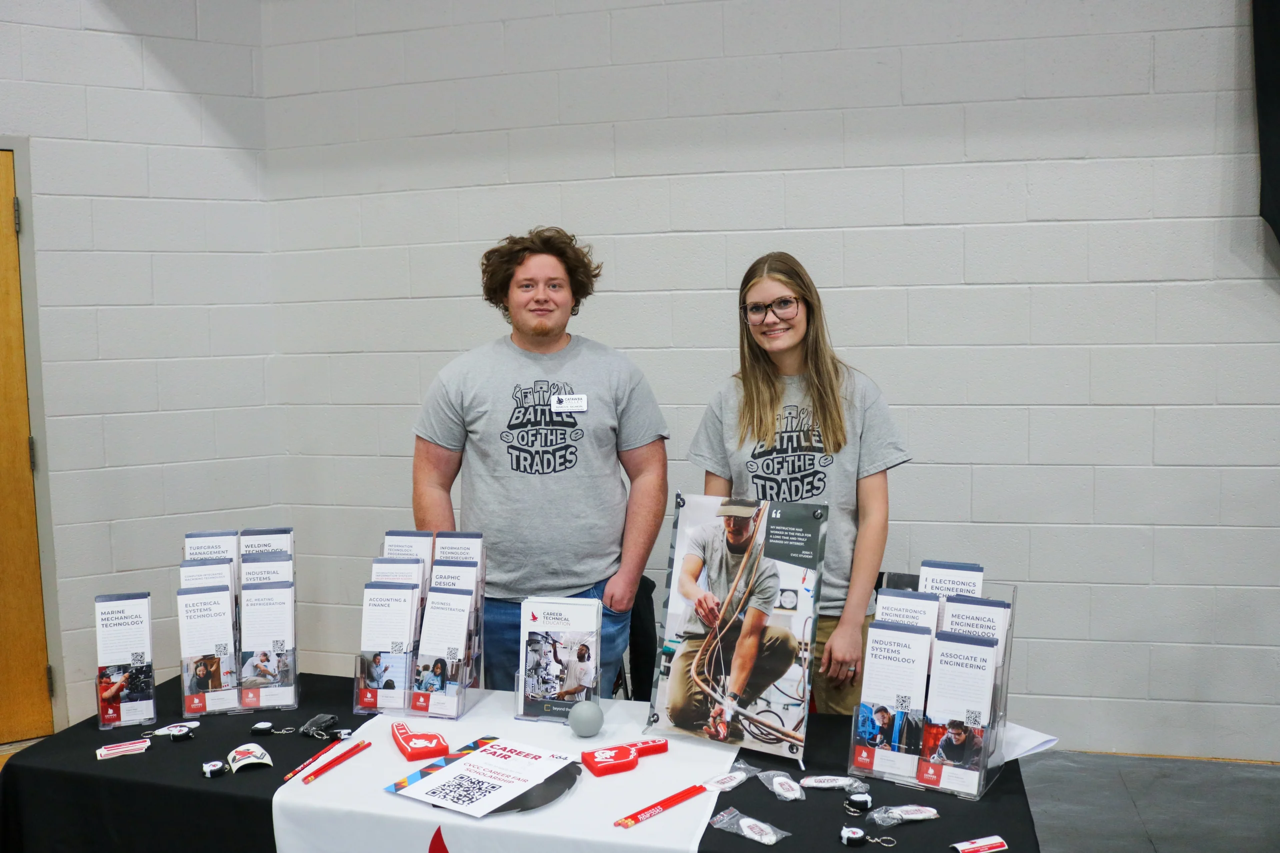 Marcus and Piper stand behind an information table with career program brochures and promotional items at a career fair.