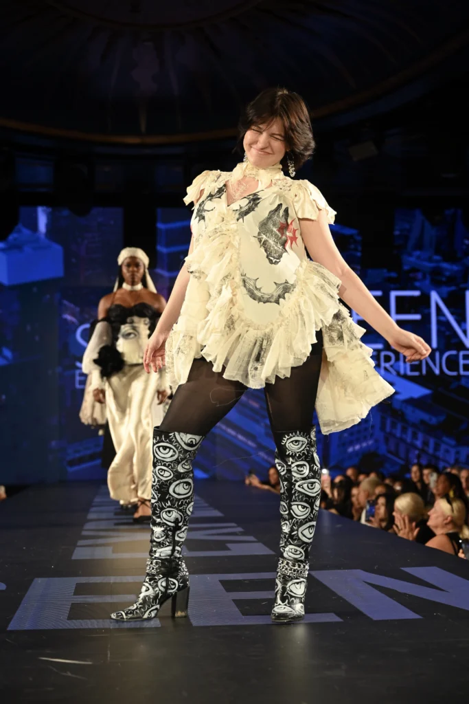 Tyler from Tyler By Tyler Designs stands on a fashion runway wearing a flowing cream ruffled top with graphic details, black leggings, and tall black‑and‑white boots featuring eye designs, while another model walks behind them.