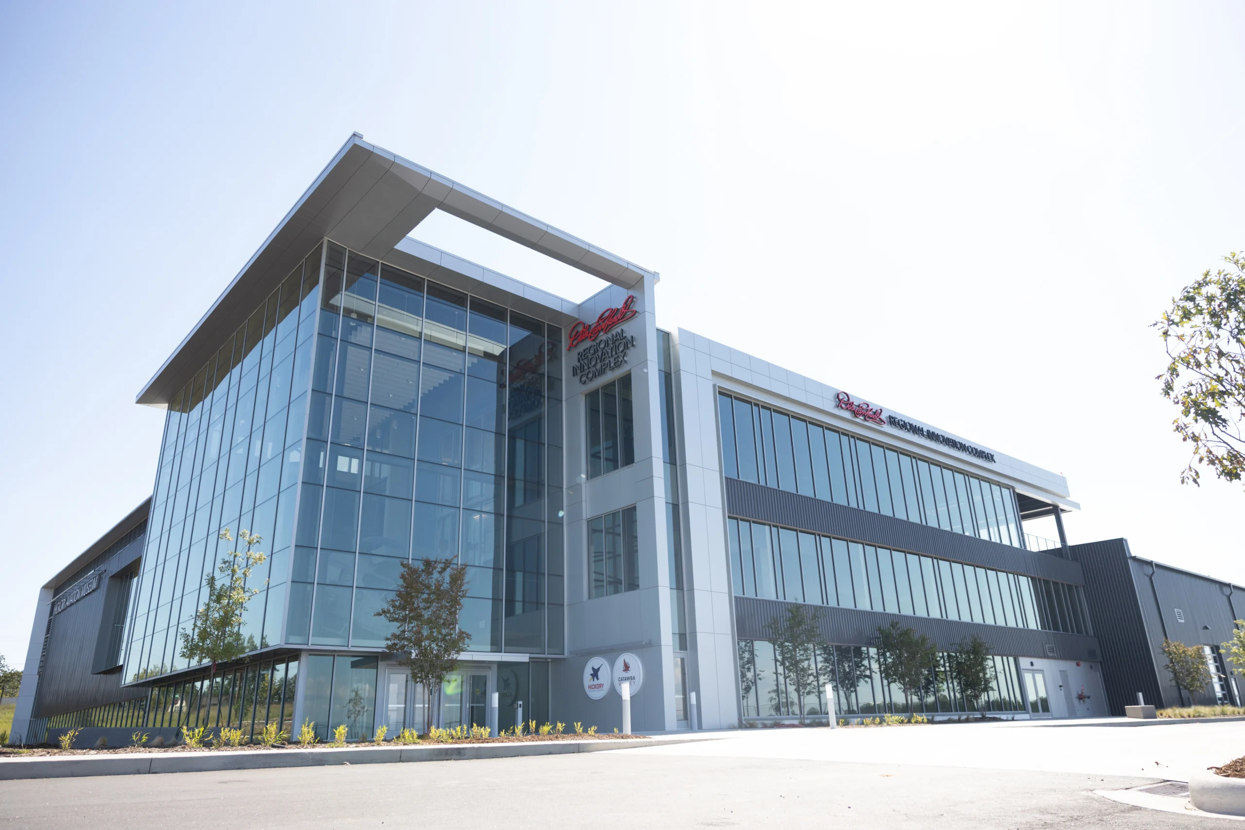 The Dale Earnhardt Regional Innovation Complex, a modern glass-and-steel building under a clear sky, features large windows, minimal landscaping, and reflective surfaces that emphasize its sleek, contemporary design.